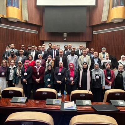 A group of people pose for a group photo inside a large conference room with wooden paneling and empty seats in the foreground.