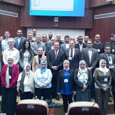 A large group of professionals, both men and women, pose together for a group photo in a conference room with a large screen behind them.