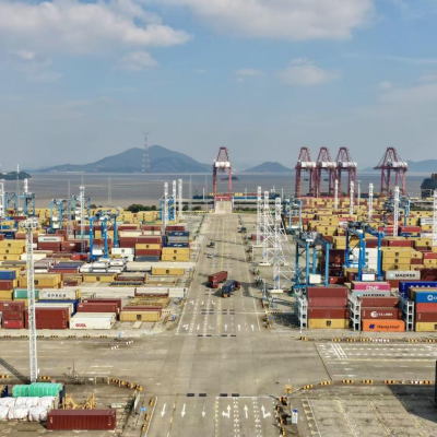 Aerial view of a large shipping port with stacked cargo containers, cranes, trucks, and distant water and hills under a partly cloudy sky.