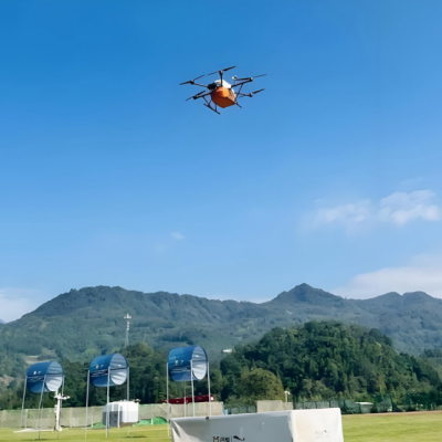 An orange drone with multiple rotors flies over a grassy field with distant hills and blue sky in the background.