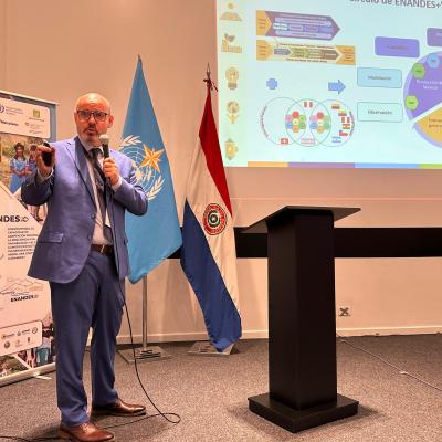 A man in a blue suit speaks into a microphone, standing next to banners, flags, and a screen displaying a colorful presentation slide.