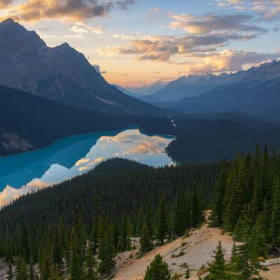 A turquoise lake winds through a forested valley surrounded by mountains at sunset, with clouds and sky reflected on the water's surface.