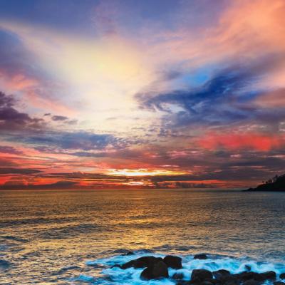 Colorful sunset over the ocean with waves crashing against rocks in the foreground and partly cloudy sky filled with vibrant hues of orange, pink, blue, and purple.