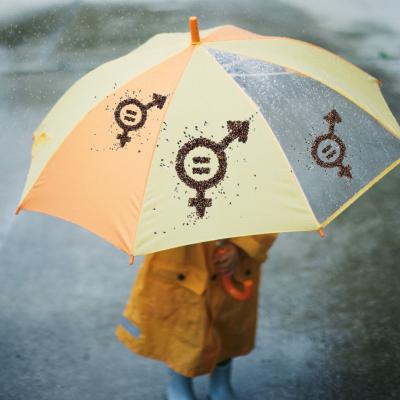 A child in a yellow raincoat stands under a yellow and orange umbrella with gender symbol designs on a rainy day.