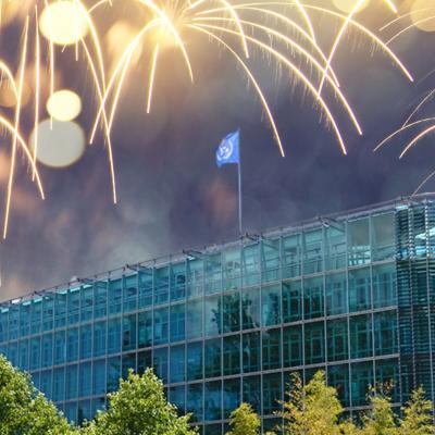 A modern glass office building with a blue flag on the roof, surrounded by trees, with fireworks lighting up the night sky above.