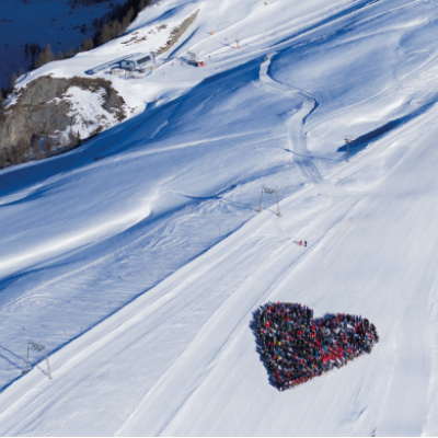 Aerial view of a snowy landscape where a group of people is gathered in the shape of a heart on the snow.