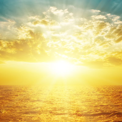 A bright golden sun rises or sets over the ocean, illuminating clouds and reflecting light on the water under a partly cloudy sky.