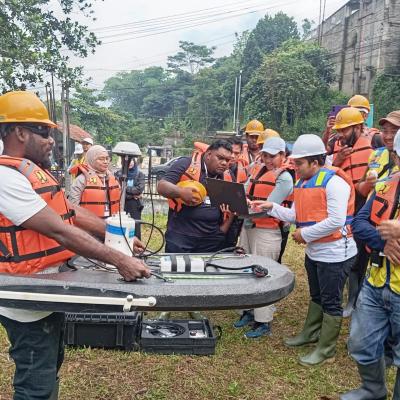 A group of people wearing safety helmets and life vests gather outdoors around equipment, with one person using a laptop and another handling a survey instrument.
