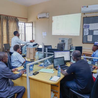Several people work at computers in an office while one person stands at the front presenting information projected onto a screen.