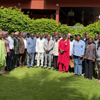 A large group of adults stands together outside on a lawn in front of a building and greenery, posing for a group photo in daylight.