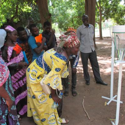 A group of people observes a demonstration of a water collection device outdoors under trees, with a woman in the foreground leaning in for a closer look.