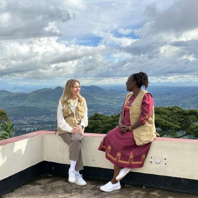 Two women sit on a low wall overlooking a scenic mountain landscape under a cloudy sky. Both wear beige vests; one is in a dress, the other in pants and a jacket.
