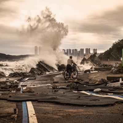 A person with a bicycle stands on a damaged coastal road, watching large waves crash onto rocks; debris and high-rise buildings are visible in the background.