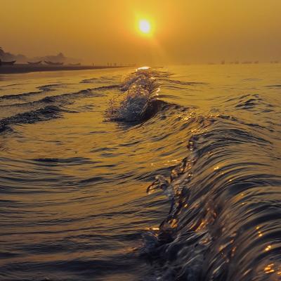 Golden sunlight reflects off gentle ocean waves near the shore at sunrise. Boats and silhouettes are visible in the distance under an orange sky.