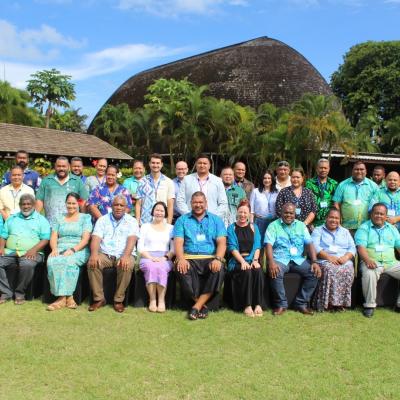 A large group of adults poses for a photo on grass in front of a tropical building with thatched roof and palm trees, under a partly cloudy sky.
