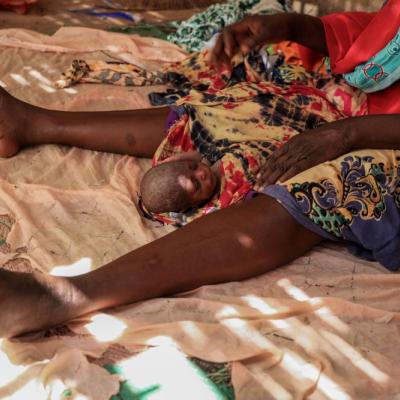 A newborn baby lies on a cloth between an adult woman's legs, with both resting on a patterned fabric floor.
