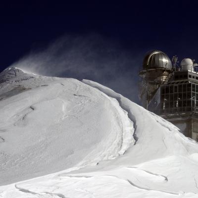 Snow-covered peak with wind-blown snow in the foreground and a building with a dome observatory structure in the background against a dark sky.