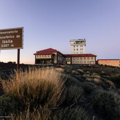 A large building complex sits on a rocky hillside at sunset, with a sign reading "Observatorio Atmosférico de Izaña 2367 m" in the foreground.