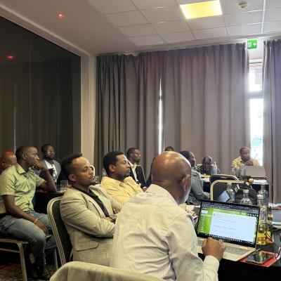 A group of people seated at tables in a conference room listen to a man presenting near a screen and a display banner. Laptops and bottles are on the tables.