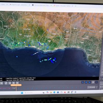 A computer monitor displays a weather radar interface showing precipitation data over a coastal region, with various control panels and map layers visible on the screen.