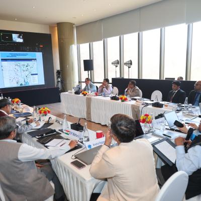 A group of people in formal attire sit around a U-shaped table with laptops and documents, viewing a large screen displaying a map and data in a modern conference room.