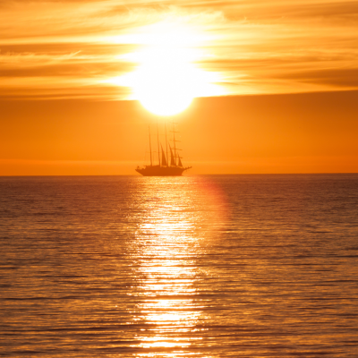 A tall ship sails on calm water at sunset, silhouetted against the bright orange sky with the sun low on the horizon and its reflection on the sea.