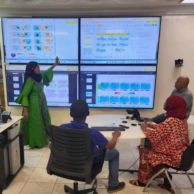Four people in an office look at large screens displaying maps and data charts; one woman stands and points to the screen while others sit and observe.