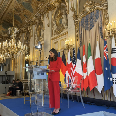 A woman in a red suit speaks at a clear podium in an ornate room with chandeliers and multiple national flags displayed behind her.