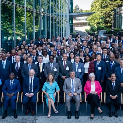 A large group of formally dressed people pose for a group photo outside a modern building, flanked by two United Nations flags.