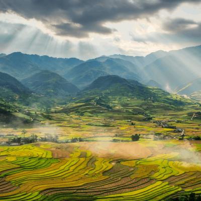 Sunlight streams through clouds over terraced rice fields and green mountains, creating a patchwork landscape in a rural valley.