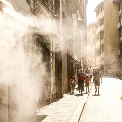 People walk down a sunlit street as mist or steam sprays from a building on the left, creating a hazy effect in the air.