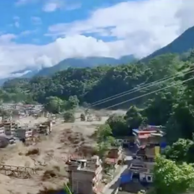 A flooded river runs through a town in a mountainous area; damaged buildings and debris are visible. A red cross with Nepali text appears in the top right corner.