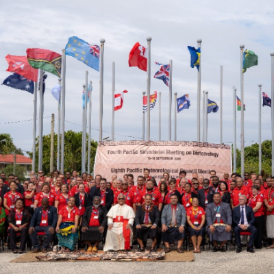 A large group of people pose for a photo in front of international flags and a banner for the Pacific Ministerial Meeting on Meteorology.