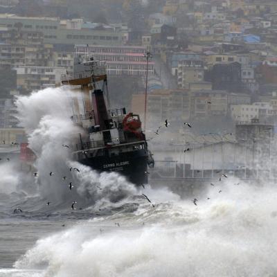 A ship being struck by large waves in a harbor, with a hillside city in the background.