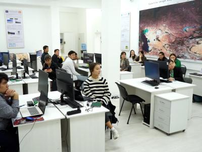 A classroom with several students sitting at desks with computers. They are attentively facing the front where an instructor is visible. A large, detailed map is displayed on the wall.