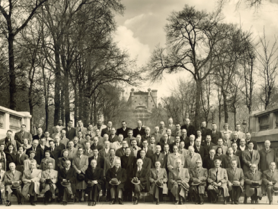 A large group of men in suits and coats pose for a formal photo outdoors, with trees lining a path and a building visible in the background.