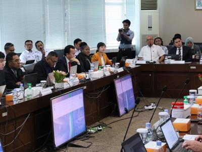 A large group of people in a conference room sit around a U-shaped table with microphones, laptops, and papers. A man stands at the back with a camera. Screens display information.