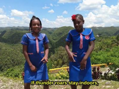 Two girls in blue school uniforms stand outdoors on a hilltop with green hills in the background. Subtitle reads, "If thunder roars stay indoors," in yellow text.