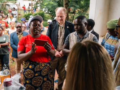 A woman in a red shirt speaks to a group of people gathered outdoors, some wearing badges and vests, while others observe in the background.