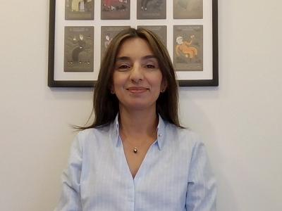 A woman with straight brown hair wearing a light blue shirt sits in front of a white wall with a framed artwork featuring several illustrations.