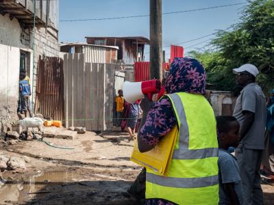 A person in a yellow vest holds a megaphone and documents while addressing people in an informal settlement area with scattered debris and makeshift structures.