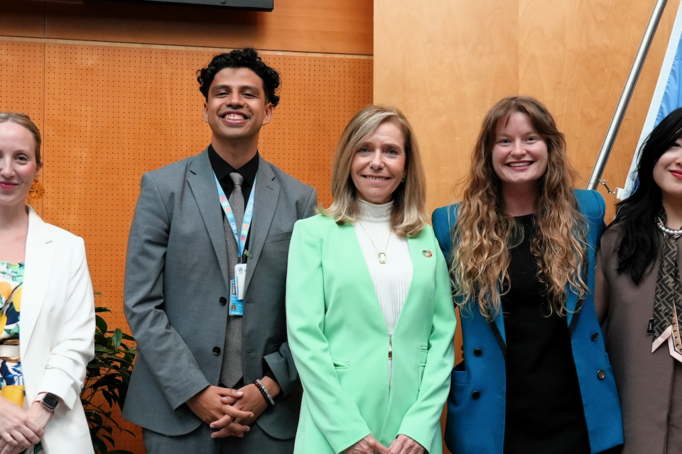 Five people are standing side by side in front of a wooden paneled wall and a flag, smiling for a group photo.