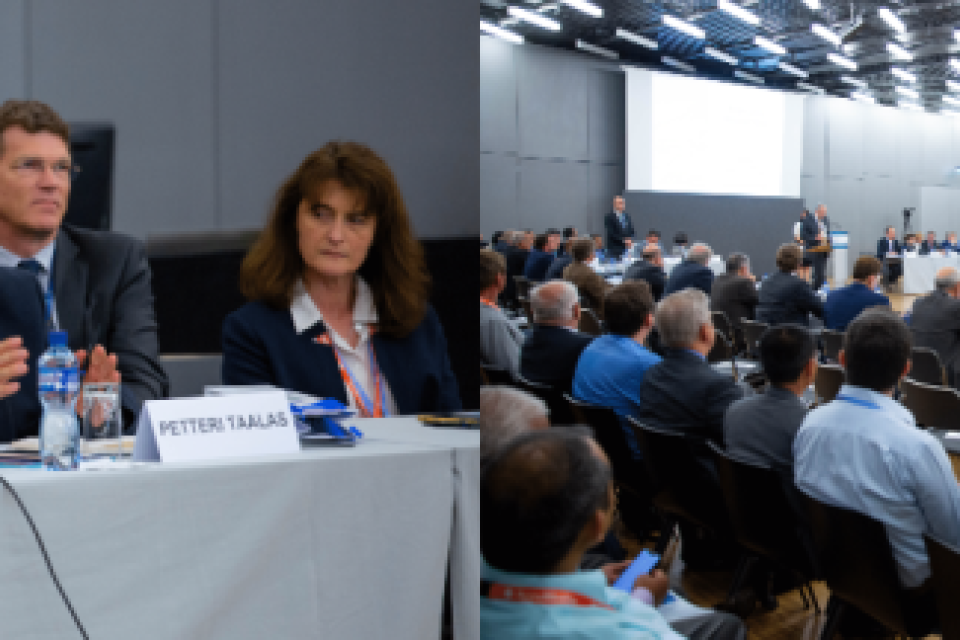 Wide-angle view of a conference room with a panel of speakers and a large audience. Participants sit at a rectangular arrangement of tables with microphones and nameplates.