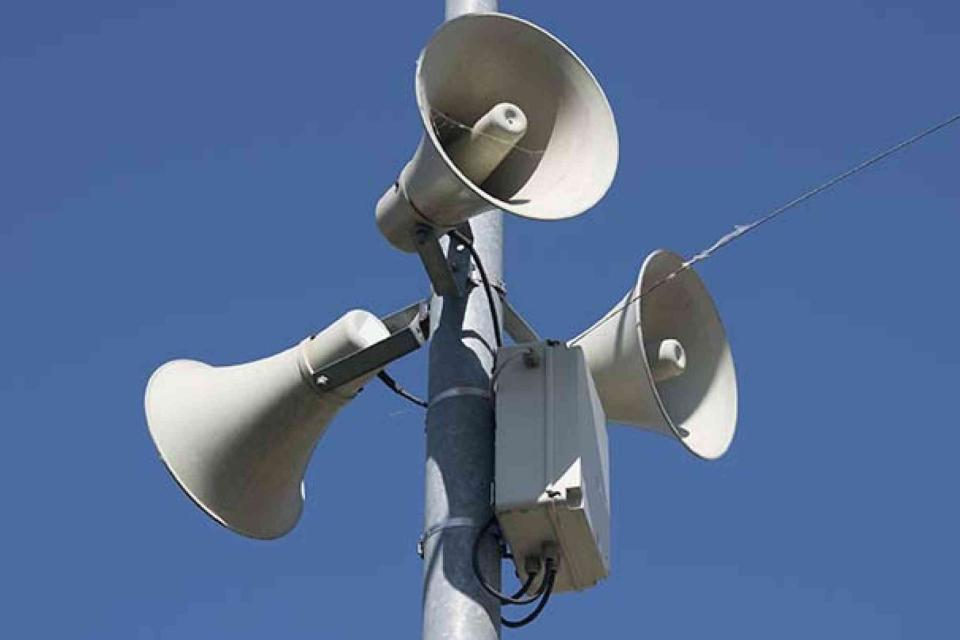 Three loudspeakers mounted on a metal pole against a clear blue sky.