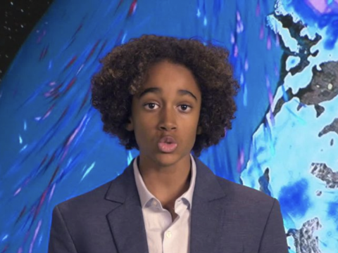 A young person with curly hair, wearing a suit, in front of a blue-toned map projection.